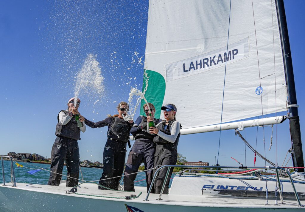 Four sailors celebrate on a sailboat by spraying champagne, with the name “LAHRKAMP” visible on the sail.