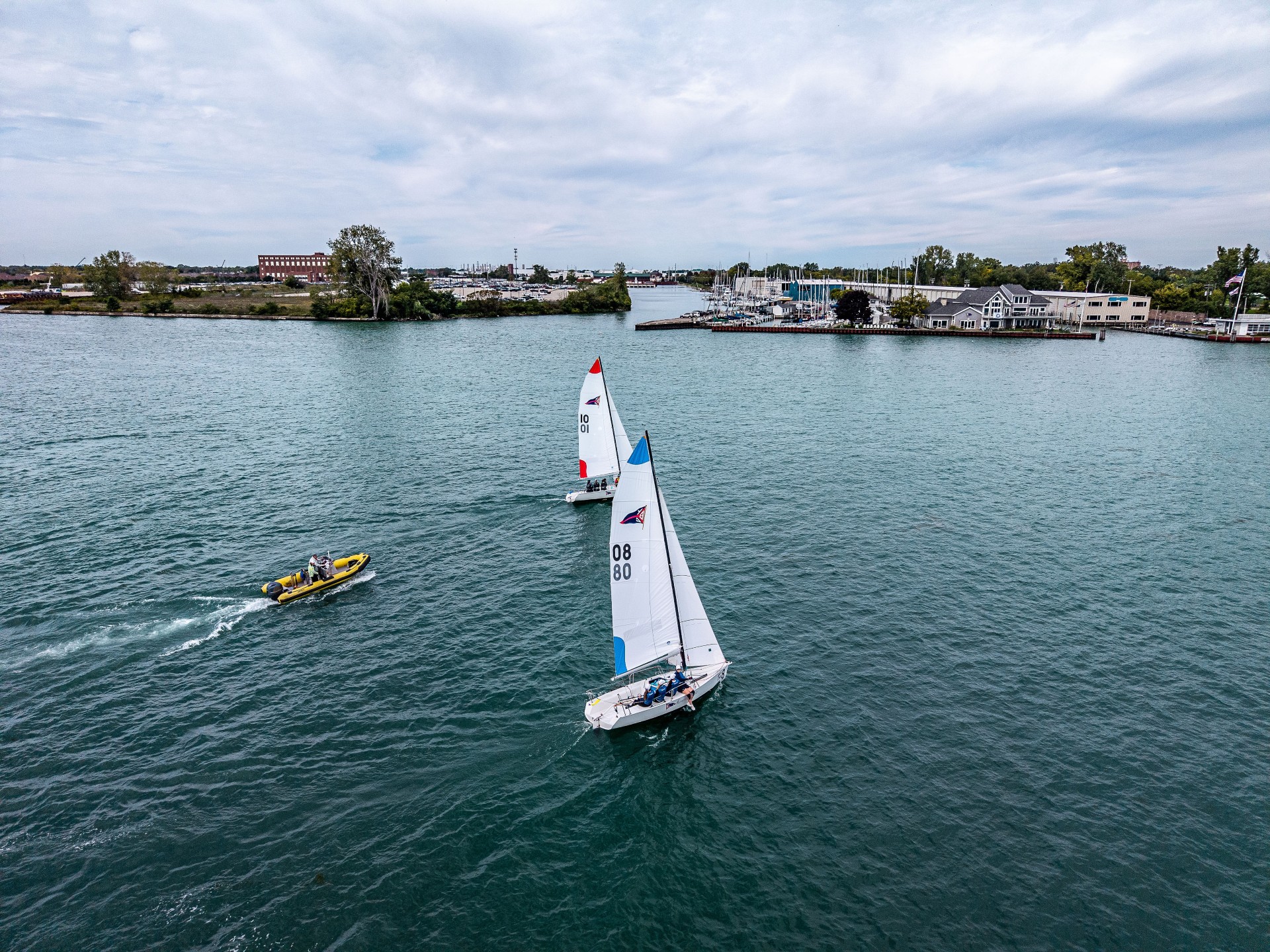 Two sailboats racing on a harbor with a yellow safety boat nearby.