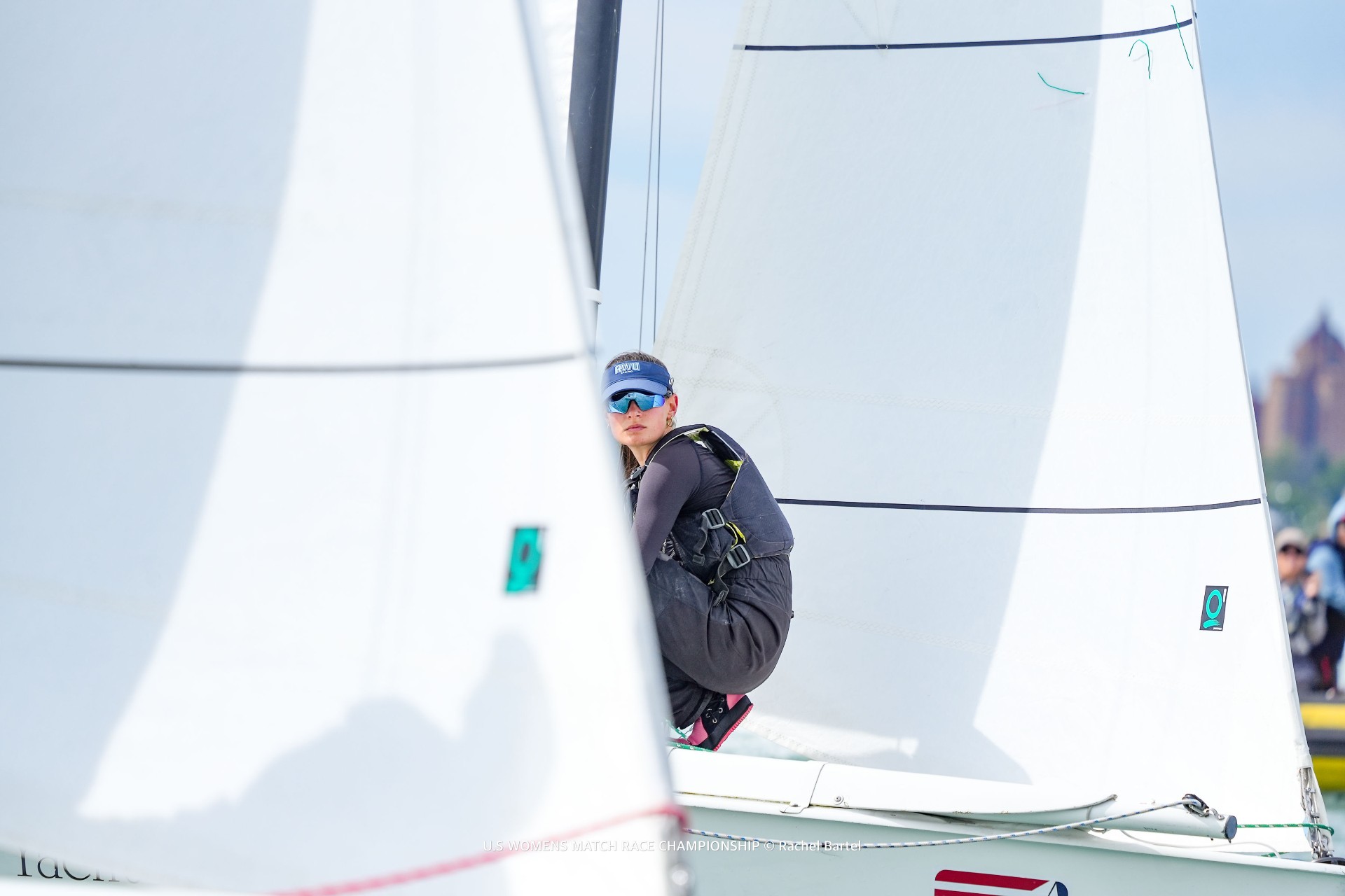 A sailor wearing a life vest and sunglasses crouches on the deck of a sailboat between two large sails during a race.