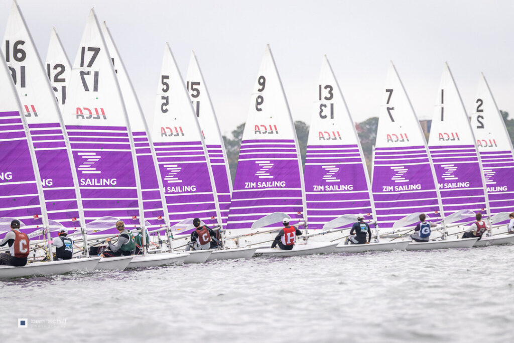 A fleet of small sailboats with purple and white sails featuring the “Zim Sailing” logo racing on the water.