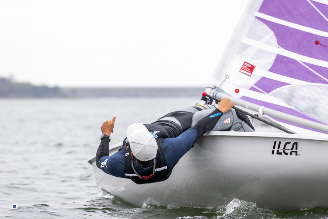 A sailor in a white cap and wetsuit leans far over the side of a small sailboat with a purple and white sail, giving a thumbs-up.