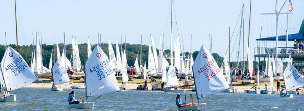 Small sailboats with white sails marked “A2U USA” navigate on the water in front of a busy sandy beach lined with many other moored boats.