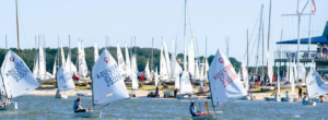 Small sailboats with white sails marked “A2U USA” navigate on the water in front of a busy sandy beach lined with many other moored boats.